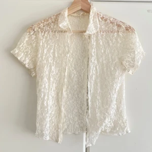 Vit spetsblus med kort ärm - Säljer en superfin vit blus i spets med korta ärmar och klassisk krage. Blusen är genombruten och har ett romantiskt uttryck, perfekt att styla lager på lager. Materialet är lätt och luftigt, vilket gör den extra bekväm.