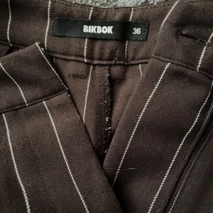 Bruna randiga kostymbyxor - Snygga bruna kostymbyxor från BikBok i storlek 36. Byxorna har vita vertikala ränder och en rak, lite loose passform. Perfekta för en clean och trendig look. Materialet känns mjukt och bekvämt, och midjan är normalhög till hög.