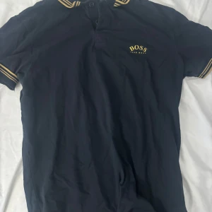 Marinblå BOSS t-shirt med gula detaljer - Snygg svart t-shirt från BOSS med guld logga på bröstet och guld ränder på krage och ärmslut. Klassisk passform och korta ärmar, perfekt för en chill och stilren look. Materialet känns mjukt och skönt mot huden.