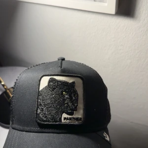 Svart keps från Goorin Bros Panther - Snygg svart keps från Goorin Bros med broderad panterpatch framtill. Kepsen har mesh på baksidan för extra ventilation och justerbar passform. Märkt med 'THE FARM' på insidan. Perfekt för dig som gillar djurmotiv och streetstyle.