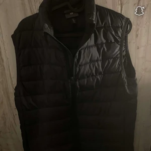 Svart dunväst från H&M, storlek S - Säljer en svart dunväst från H&M i storlek S. Västen är quiltad med horisontella sömmar, har hög krage och dragkedja framtill. Perfekt att slänga över en hoodie för extra värme. Enkel och stilren design som funkar till det mesta.