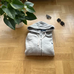 Grå hoodie från Polo Ralph Lauren - Snygg ljusgrå hoodie från Polo Ralph Lauren med dragkedja och huva. Klassisk logga broderad i mörkblått på bröstet. Tröjan har snörning i huvan, långa ärmar och är tillverkad i mjuk bomull. Perfekt för en chill och stilren look.