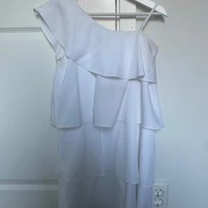 Vit omlottklänning med volanger - Superfin vit klänning med asymmetrisk axel och volangdetaljer i flera lager. Klänningen har en axelbandslös sida och en smal axelrem, vilket ger en modern och trendig look. Använd 1 gång på skolavslutningen. Har en fläck där fram men det är inget som inte försvinner i tvätten🥰