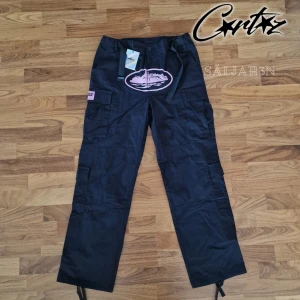 Corteiz Cargobyxor - Säljer helt nya Corteiz Guerillaz Cargo Pants i svart med rosa detaljer, originaletiketter kvar. Storlek: L (passar mer som M). Material: Slitstarkt ripstop-tyg, bekvämt. Design: Broderade & tryckta detaljer, cargofickor med lock, justerbara band i midja & benslut. Perfekt för dig som gillar exklusiv streetwear! Limited release – svåra att få tag på! 📦 Snabb leverans – skickas samma dag! 📦 Kontakta mig vid intresse eller funderingar!📌