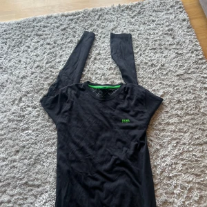 Svart långärmad tröja från Hugo Boss - Storleken kan va mindre än XXL för att tröjan är köpt för så länge sen, den gröna detaljen ska vara så den va köpt så. Svart långärmad tröja från Hugo Boss med grön logga på bröstet och coolt grönt band i halsen. Tröjan har rund halsringning och är tillverkad i mjuk bomull, perfekt för en clean och stilren look.