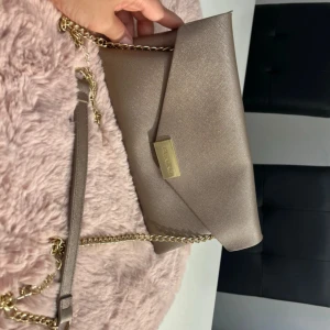 Beige kuvertväska från Valentino - Stilren beige kuvertväska från Valentino med guldfärgad kedja och magnetlås. Väskan har en elegant, slät yta och klassisk triangelformad flärp framtill. Perfekt att bära crossbody eller i handen för en chic look.