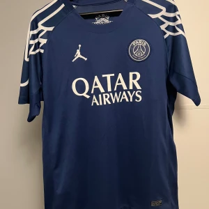 PSG blå fotbollströja Jordan - Snygg PSG matchtröja i marinblått med vita detaljer och tryck från Jordan och Qatar Airways. Kortärmad modell med coola vita mönster på axlarna. Perfekt för dig som vill ha en ikonisk look på planen eller läktaren.