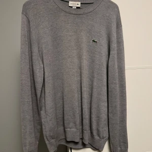 Grå stickad tröja från Lacoste - Snygg grå stickad tröja från Lacoste med klassisk rund halsringning och den ikoniska gröna krokodilloggan på bröstet. Tröjan har lång ärm och ribbade muddar vid ärmslut och nederkant. Perfekt för lager-på-lager och stilren look.