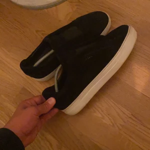 Svarta mockasneakers med vit sula - Stilrena svarta sneakers i mocka med vit platt sula. Skorna har minimalistisk design utan synlig snörning och en rund tå. Perfekta för dig som gillar en clean look och vill ha något som funkar till det mesta.