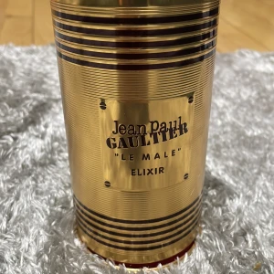 Jean Paul Gaultier Le Male Elixir - Exklusiv parfym i metallisk guldflaska formad som en manlig torso med svarta ränder. Kommer i en matchande guldfärgad burk med eleganta detaljer och röd sammetsbas. En riktig statement-doft för dig som gillar lyxig design.