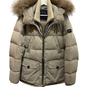 Beige dunjacka från Paul & Shark - Snygg beige dunjacka från Paul & Shark med fluffig pälskant på huvan och broderad logga på ärmen. Jackan har dragkedja, stora fickor med knappar och en mysig krage i teddy. Perfekt för kalla dagar när du vill hålla stilen. Inköpt på NK i Stockholm för 10998kr. Pälsen går att ta av samt innervästen.