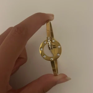 Guldigt Edblad armband med stenar - Snyggt armband från Edblad i guldtonad metall med en rund detalj framtill, dekorerad med glittriga stenar. Armbandet har en stilren och modern design som verkligen sticker ut. Perfekt för dig som gillar att addera lite extra bling till din look.