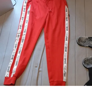 Röda mjukisbyxor Plein Sport - Riktigt snygga röda mjukisbyxor från Plein Sport med vita sidoränder där det står Plein Sport i svart. Byxorna har dragsko i midjan och muddar vid bensluten. Perfekta för chill eller träning, med en skön och sportig vibe.