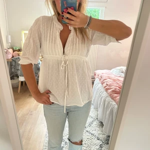 Vit blus med prickar - Supersöt vit blus med diskreta prickar, knytband i midjan och lätt rynkade detaljer. Blusen har v-ringning, trekvartsärmar och volangkant vid halsen. Perfekt för en fräsch och luftig look till sommaren eller våren.