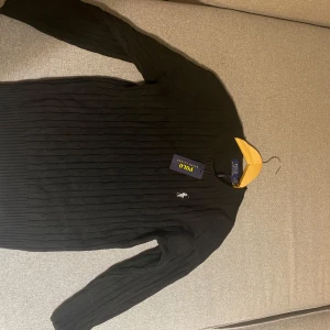 Svart stickad tröja Polo Ralph Lauren (storlek S) - Svart stickad tröja från Polo Ralph Lauren med klassisk rund halsringning och diskret broderad logga på bröstet. Tröjan har lång ärm och ribbade muddar, perfekt för en stilren och enkel look.