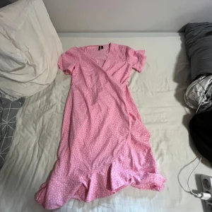 Rosa prickig omlottklänning Vero Moda - Supersöt rosa omlottklänning från Vero Moda med vita prickar, volangkant och knyt i sidan. Klänningen har korta ärmar och v-ringad hals. Perfekt för sommarens alla tillfällen och ger en fräsch och lekfull vibe. Materialet är mjukt och lätt, troligtvis polyester.