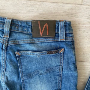 Blå slim fit jeans från Nudie Jeans - Säljer ett par blå skinny jeans från Nudie Jeans med klassisk femficksdesign och slitna detaljer. Jeansen har en smal passform och är tillverkade i mjukt denimtyg med orangea sömmar och Nudie-logga på bakfickan. Perfekt för dig som gillar en cool och avslappnad stil.