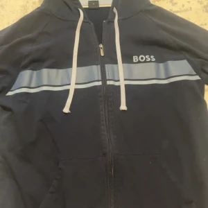 Marinblå hoodie från BOSS med dragkedja - Säljer en marinblå hoodie från BOSS i storlek M. Tröjan har hel dragkedja, huva med vita snören och en ljusblå rand över bröstet. Praktiska fickor framtill och klassisk BOSS-logga på bröstet. pris kan diskuteras!