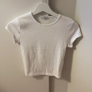 Vit ribbad topp från Bershka - Ett jättebra basplagg. En stilren vit ribbad topp från Bershka i kortärmad modell. Toppen har rund halsringning och är figurnära med en croppad känsla. Perfekt att matcha med jeans eller kjol för en clean look.