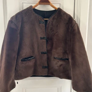 Brun pälsjacka med knäppning - Mysig brun pälsjacka med raka linjer och snygga detaljer i svart skinn vid knäppning och fickor. Jackan är croppad och har lång ärm, perfekt för kalla dagar när du vill ha en cool och trendig look. En av knapparna har lossnat, men jag har kvar knappen så det är lätt att sy på igen. Inte hundra på storlek, men skulle säga att den passar xs-m beroende på passform.