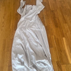 Ljusgrå långklänning med kort ärm - En stilren ljusgrå långklänning med korta ärmar och enkel, rak passform. Klänningen har en mjuk och flowy känsla, perfekt för dig som gillar minimalistisk stil. Materialet ser ut att vara bomull eller polyester, vilket gör den bekväm och lätt att bära.