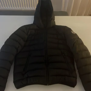 Svart pufferjacka från Colmar - Säljer en svart pufferjacka från Colmar Originals med huva och dragkedja framtill. Jackan har quiltad design och två sidofickor med dragkedja. Perfekt för kyliga dagar och riktigt snygg till streetwear-stilen.