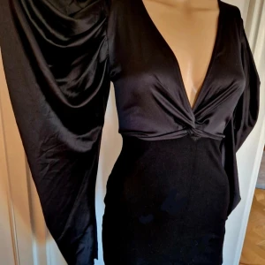 Svart festklänning från Zara - Supersnygg svart klänning från Zara med djup v-ringning och twistad detalj framtill. Klänningen har långa, vida ärmar i satinliknande material och figurnära passform  i kjol med stretch i.Perfekt för dig som vill sticka ut med en elegant och trendig look.Stl på etiketten är S men är väldigt liten.Dockan är i stl 36/s och det sitter mkt tajt på denne