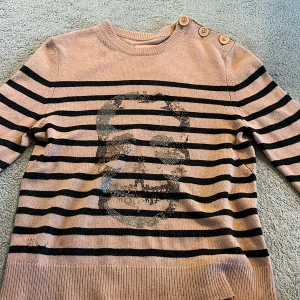 Randig tröja med döskalle från Zadig & Voltaire - ÄKTA!! Cool stickad tröja från Zadig & Voltaire i beige med svarta ränder och glittrig döskalle framtill. Tröjan har rund hals, tre knappar på axeln och långa ärmar.