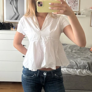 Vit blus med v-ringning och kort ärm - Säljer en söt vit blus med v-ringning och korta ärmar. Modellen är luftig och har en lätt utställd, flowy passform som ger en avslappnad vibe. Perfekt till jeans eller kjol för en fräsch look. Gjord i bomull för en skön känsla.