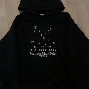 Maison Margiela svart hoodie - Svart hoodie från Maison Margiela med ikoniskt siffertryck och logga på bröstet. Klassisk huva och lång ärm, tillverkad i mjuk bomull. Perfekt för dig som gillar streetstyle och vill sticka ut med en exklusiv look.S som passar xs
