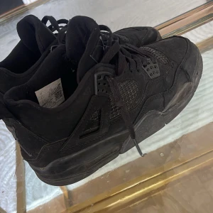 Svarta Nike Air Jordan 4 sneakers - Säljer ett par helsvarta Nike Air Jordan 4 sneakers med klassisk mesh och detaljer i mocka. Skorna har snörning, rund tå och den ikoniska Jumpman-loggan på hälen och plösen. Perfekta för dig som gillar streetstyle och vill ha en clean look.