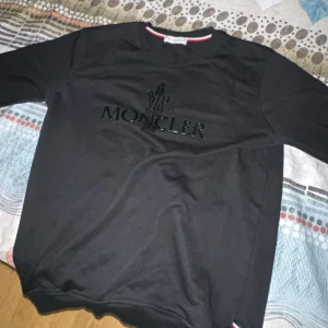 Svart Moncler sweatshirt  - Svart sweatshirt från Moncler med stort, svart Moncler-tryck på bröstet och en snygg patch på ärmen. Tröjan har rund hals och långa ärmar, tillverkad i mjuk bomull. Perfekt för dig som gillar stilrena designer och exklusiva märken.