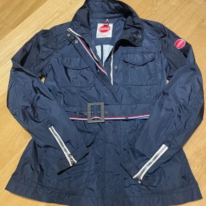 Mörkblå vindjacka från Colmar - Snygg mörkblå vindjacka från Colmar med bälte i midjan och coola detaljer som dragkedjor vid ärmsluten. Jackan har flera fickor framtill och en sportig känsla med röd, vit och blå rand på bältet.  Storlek motsvarar S. 