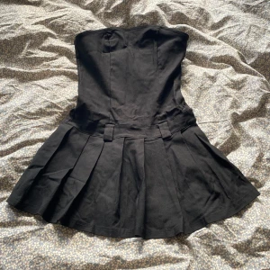 Svart axelbandslös kort klänning - Säljer en svart axelbandslös klänning med kort längd och plisserad kjol. Klänningen har en figurnära överdel och breda hällor i midjan, vilket ger en cool och edgy vibe. Perfekt för dig som gillar minimalistisk stil med en twist. Har även undershorts.