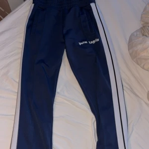 Palm Angels blå trackpants S - Säljer ett par blå trackpants från Palm Angels med vita sidoränder och broderad logga på ena benet. Byxorna har elastisk midja, raka ben och är gjorda i polyester för en sportig vibe. Perfekta för dig som gillar streetwear och snygga detaljer.