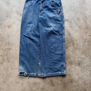 JNCO Jeans lång jeanskjol med patch - Säljer en blå lång jeanskjol från JNCO Jeans med coola broderade patches fram och bak. Kjolen har klassiska fickor, rå kant nedtill och loose passform. Perfekt för dig som gillar streetstyle och vill sticka ut med en unik denimlook.