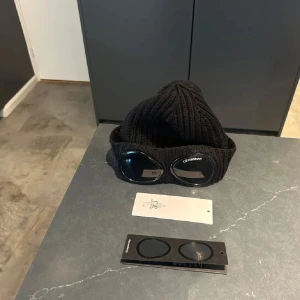 Svart Goggle-mössa från C.P. Company - Snygg svart stickad mössa från C.P. Company med deras ikoniska glasögondetaljer framtill. Inhandlad på Nordiska kompaniet för två vintrar sen (Nypris 1500). Mössan är i ett mycket bra skick och säljs på grund av att jag inte använt den tillräckligt. Hör av er vid funderingar! 