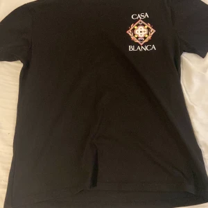 Svart Casablanca t-shirt med tryck - Svart t-shirt från Casablanca med stort färgglatt emblem-tryck och texten 'CASABLANCA' på både bröstet och ryggen. Klassisk passform och rund hals. Perfekt för dig som gillar streetwear med en exklusiv touch.