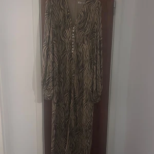 Adoore Positano långklänning zebra - Säljer en snygg Positano långklänning från Adoore med zebramönster i beige och svart. Klänningen har v-ringning, knappar framtill och långa ärmar med lätt puff. Perfekt för dig som vill sticka ut med ett coolt mönster och skön passform. Nypris 1695. Helt oanvänd med lappen kvar. 