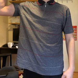 Grå piké med dragkedja och ränder - Säljer en grå pikétröja med tunna röda och vita ränder. Tröjan har korta ärmar, krage med dragkedja och en sportig look. Perfekt för dig som gillar stilrena och bekväma plagg. Liten för att vara large så funkar för m, möjligtvis s 