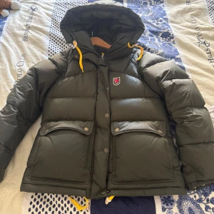 Grön dunjacka från Fjällräven Expedition Series - Säljer en mörkgrön dunjacka från Fjällräven Expedition Series med huva och gula detaljer. Jackan har två stora fickor framtill, knappar och dragkedja. Perfekt för kalla dagar och riktigt varm.  Jackan köpte jag för 5 månader sedan, köptes för 7000 kr, säljer för 5000, men priset kan diskuteras.