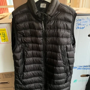 Svart dunväst från C.P. Company - Jag säljer min svarta Cp company väst. Det är ett mini hål på den men den är lagad med en svartklädd lapp så man inte ens märker den, fler bilder kan fås vid förfrågan och priset kan ändras vid snabb affär 