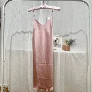 Ljusrosa satinklänning med spetsdetaljer - Säljer en ljusrosa långklänning i satin med tunna axelband och v-ringad hals. Klänningen har fina spetsdetaljer längs ryggen och axelbanden är korsade i ryggen för en extra snygg look. Perfekt för sommarens tillfällen!