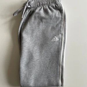 Grå mjukisbyxor från Adidas - Snygga grå mjukisbyxor från Adidas med vita ränder längs sidorna och logga på låret. Byxorna har resår och dragsko i midjan samt en bakficka. Perfekta för en avslappnad stil.