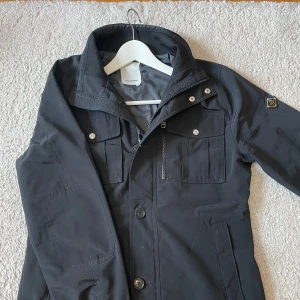  J.Lindeberg field jacket  - Säljer en svart jacka från J.Lindeberg med klassisk krage, dragkedja och knappar framtill. Jackan har två bröstfickor med lock och knappar samt två sidofickor. Jackan är i storlek M men är mer som en S. Perfekt för dig som vill ha en stilig och sjysst jacka till sommarn/våren