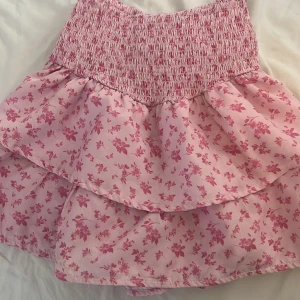 Rosa blommig volangkjol - Supersöt rosa kjol med blommigt mönster och volanger. Kjolen har en smockad midja som ger en bekväm passform och dubbla lager volang för extra volym. Perfekt för vår och sommar!