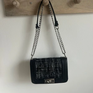 Svart axelväska med kedja och tweed - Snygg svart axelväska med silverfärgad kedja och lock i svartvitt tweedmönster. Väskan har ett stilrent spänne i metall framtill och detaljer i syntetmaterial. Perfekt för att lyfta din outfit med en trendig accessoar.