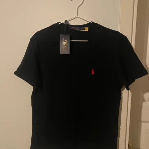 Svart t-shirt från Polo Ralph Lauren - Säljer en svart t-shirt från Polo Ralph Lauren med klassisk röd broderad logga på bröstet. T-shirten har rund halsringning och korta ärmar. Perfekt för en stilren och enkel look. Helt oanvänd, lappen sitter kvar