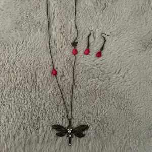 Set med halsband och örhängen med trollslända och röda droppar - •Säljer ett nytt smyckeset med ett långt halsband med trollsländehänge i antikguld och glittrande stenar samt matchande örhängen med röda droppformade hängen.  •Kedjan och detaljerna är i metall med vintagekänsla. 
