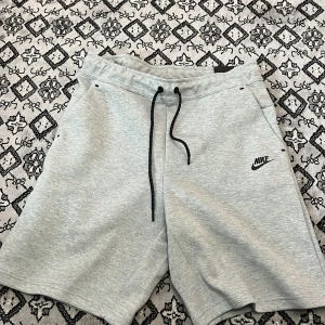 Grå shorts från Nike med snörning - Snygga ljusgrå shorts från Nike med svart logga på vänster ben. De har elastisk midja med svart snörning och två sidofickor. Perfekta för en avslappnad stil eller träning. Mjukt material som känns skönt mot huden.